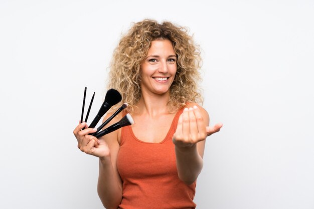 Junge Blondine mit dem gelockten Haar, das viel Make-upbürste einlädt, mit der Hand zu kommen hält. Schön, dass Sie gekommen sind