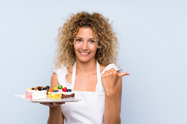 Junge blondine mit dem gelockten haar, das viele verschiedene minikuchen zeigen auf die seite hält, um ein produkt darzustellen