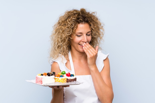 Junge Blondine mit dem gelockten Haar, das viele verschiedenen Minikuchen lächeln viel hält