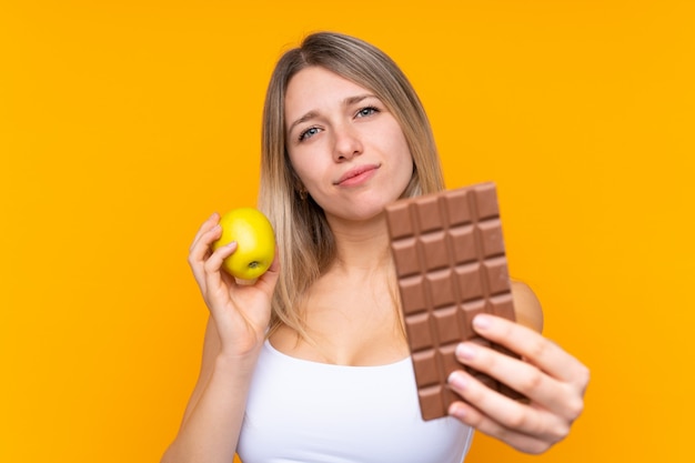 Junge Blondine über der lokalisierten blauen Wand, die eine Schokoladentablette in einer Hand und einen Apfel in der anderen nimmt