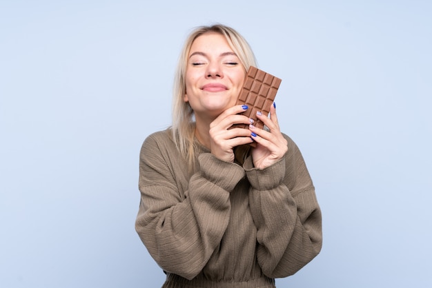 Junge Blondine über der lokalisierten blauen Wand, die eine Schokoladentablette und ein glückliches nimmt