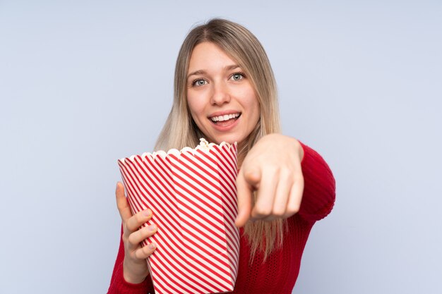 Junge Blondine über der lokalisierten blauen Wand, die einen großen Eimer Popcorn beim Zeigen von Front hält