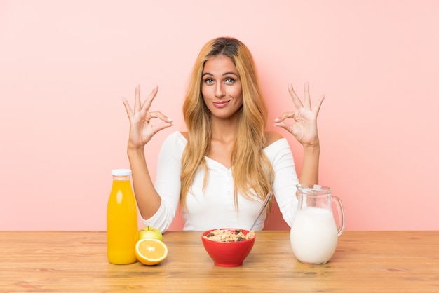 Junge Blondine, welche die Frühstücksmilch zeigt ein okayzeichen mit den Fingern essen