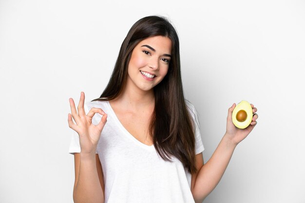 Junge Brasilianerin, die eine Avocado isoliert auf weißem Hintergrund hält und mit den Fingern ein Ok-Zeichen zeigt