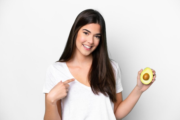 Junge Brasilianerin, die eine Avocado isoliert auf weißem Hintergrund mit überraschtem Gesichtsausdruck hält