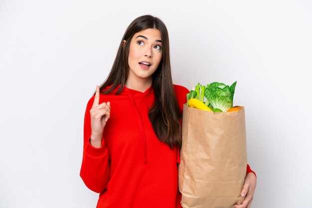 Junge Brasilianerin, die eine Lebensmitteleinkaufstasche isoliert auf weißem Hintergrund hält und an eine Idee denkt, die mit dem Finger nach oben zeigt