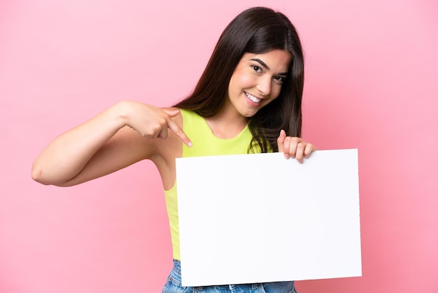 Junge Brasilianerin isoliert auf rosa Hintergrund, die ein leeres Plakat mit fröhlichem Ausdruck hält und darauf zeigt