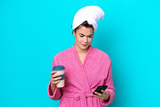 Junge Brasilianerin mit Bademantel isoliert auf blauem Hintergrund mit Kaffee zum Mitnehmen und einem Handy