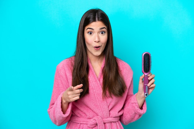 Junge Brasilianerin mit Haarbürste isoliert auf blauem Hintergrund überrascht und zeigt nach vorne