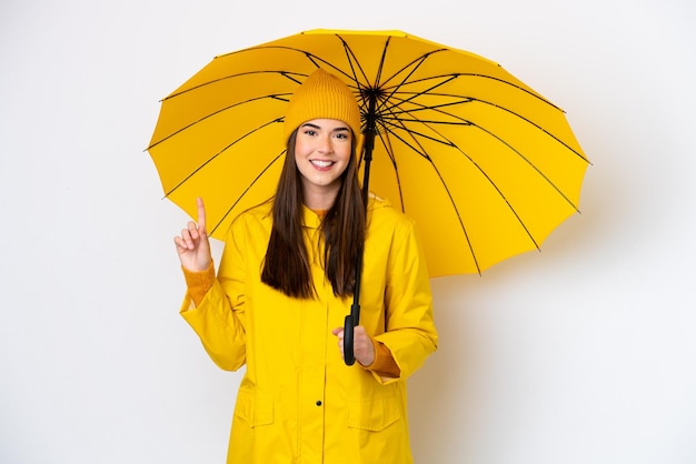 Junge Brasilianerin mit regenfestem Mantel und Regenschirm isoliert auf weißem Hintergrund, die auf eine großartige Idee zeigt