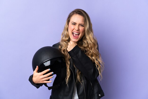 Junge brasilianische Frau, die einen Motorradhelm lokalisiert auf lila Wand hält, die einen Sieg feiert