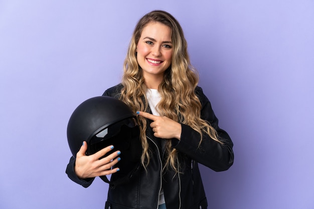 Junge brasilianische Frau, die einen Motorradhelm lokalisiert hält, der auf lila zeigt, der zur Seite zeigt, um ein Produkt zu präsentieren