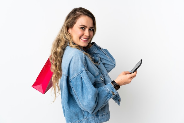 Junge brasilianische Frau lokalisiert auf weißem Hintergrund, der Einkaufstaschen hält und eine Nachricht mit ihrem Handy an einen Freund schreibt