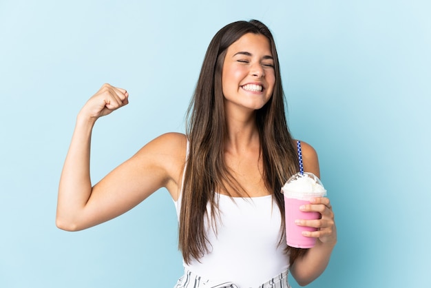 Junge brasilianische Frau mit Erdbeermilchshake auf Blau, die starke Geste tut