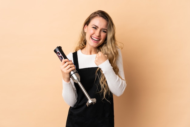 Junge brasilianische Frau mit Handmixer lokalisiert auf Beige, die einen Sieg feiert