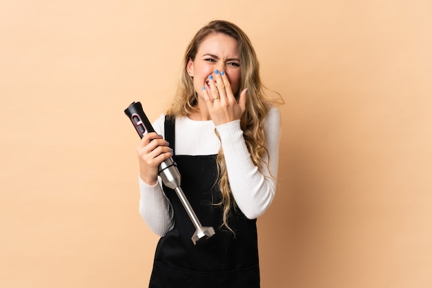 Junge brasilianische Frau mit Handmixer lokalisiert auf beige glücklich und lächelnd, das Mund mit Hand bedeckt