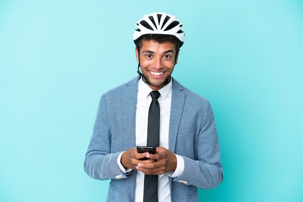 Junge brasilianische Geschäftsfrau mit Fahrradhelm auf blauem Hintergrund isoliert, die eine Nachricht mit dem Handy sendet