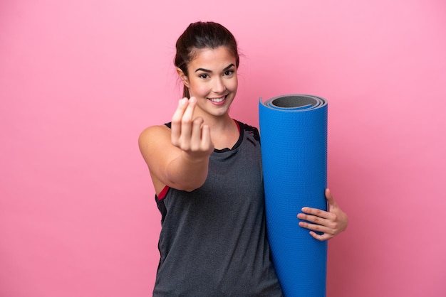 Junge brasilianische Sportlerin, die zu Yoga-Kursen geht, während sie eine Matte hält, die isoliert auf rosafarbenem Hintergrund Geld macht, Geste