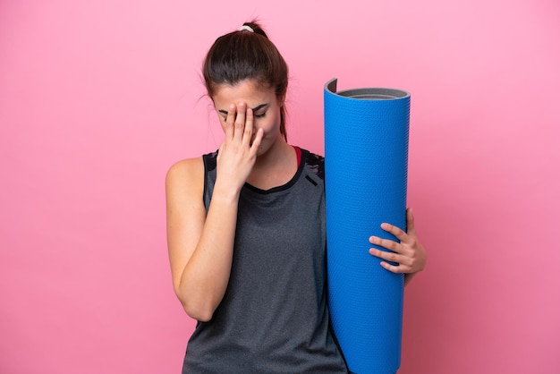 Junge brasilianische Sportlerin, die zu Yoga-Kursen geht, während sie eine Matte isoliert auf rosa Hintergrund mit müdem und krankem Ausdruck hält