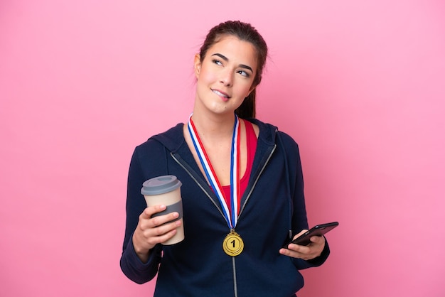 Junge brasilianische Sportlerin mit Medaillen isoliert auf rosa Hintergrund, die Kaffee zum Mitnehmen und ein Handy hält, während sie etwas denkt