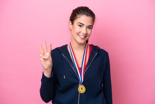 Junge brasilianische Sportlerin mit Medaillen isoliert auf rosa Hintergrund glücklich und zählt drei mit den Fingern