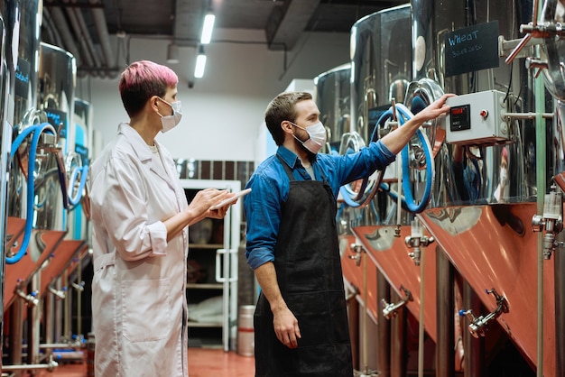 Junge Brauereiexpertin und männlicher Arbeiter in Schutzmasken, die während des Arbeitsprozesses neue Bierproduktionsanlagen testen