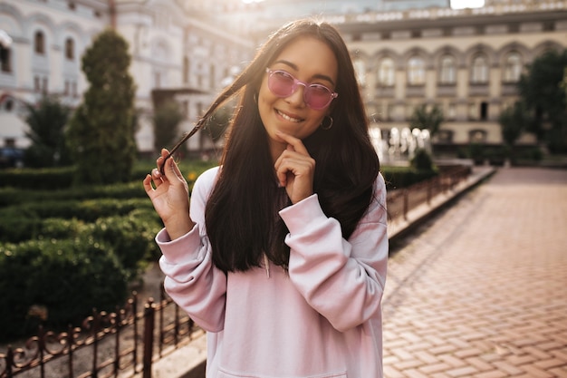 Junge brünette Asiatin in rosa Sonnenbrille und stylischem Hoodie sieht nachdenklich aus, spielt Haare und posiert draußen