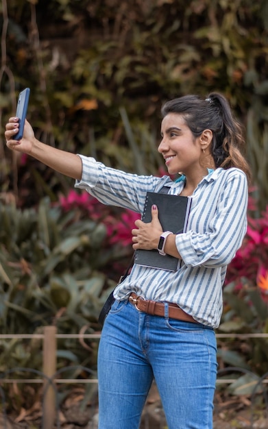 Junge brünette Frau, die ein Selfie macht, während sie ein Notizbuch außerhalb des Campus hält Konzept des College-Studenten