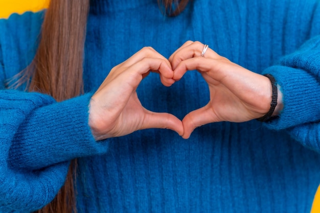 Junge brünette Frau, die einen blauen Pullover über isoliertem gelbem Hintergrund trägt und verliebt lächelt und mit den Händen ein romantisches Konzept in Form eines Herzsymbols macht