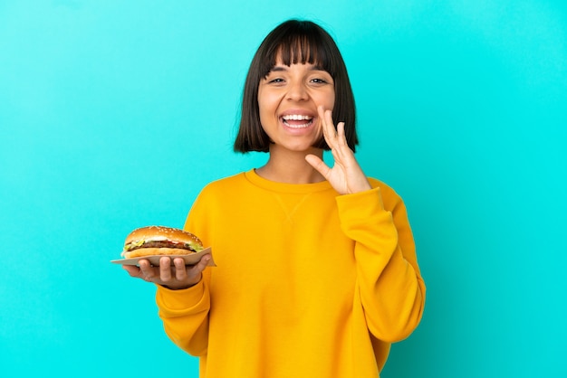 Junge brünette Frau, die einen Burger über einer isolierten Wand hält und mit weit geöffnetem Mund schreit