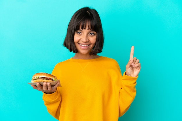 Junge brünette Frau, die einen Burger über isoliertem Hintergrund hält und auf eine großartige Idee zeigt