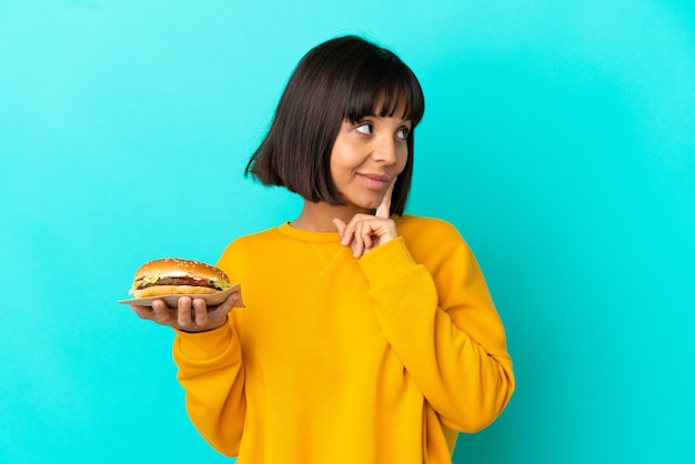 Junge brünette Frau, die einen Burger über isoliertem Hintergrund hält und eine Idee denkt, während sie nach oben schaut