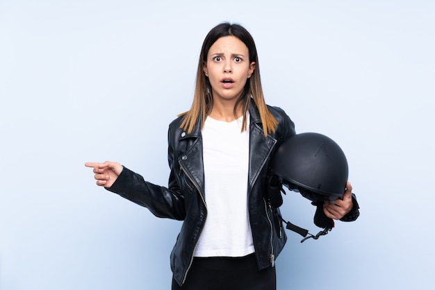 Junge brünette Frau, die einen Motorradhelm über der blauen Wand hält, überraschte und zeigende Seite