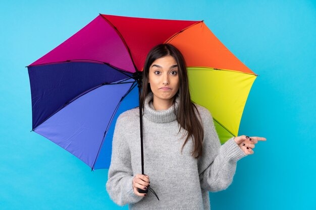 Junge brünette Frau, die einen Regenschirm über blauer Wand hält, die auf die Seiten zeigt, die Zweifel haben