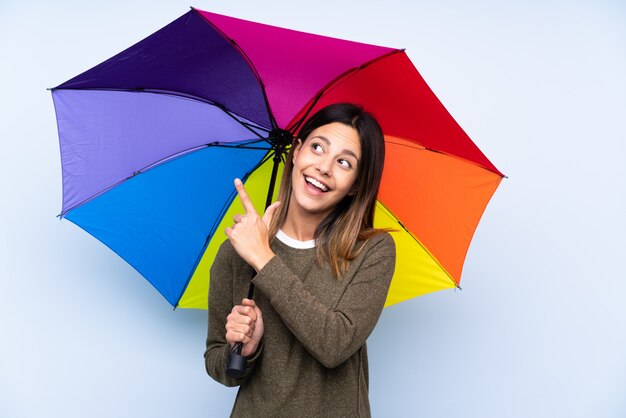 Junge brünette Frau, die einen Regenschirm über blauer Wand hält, die mit dem Zeigefinger eine große Idee zeigt