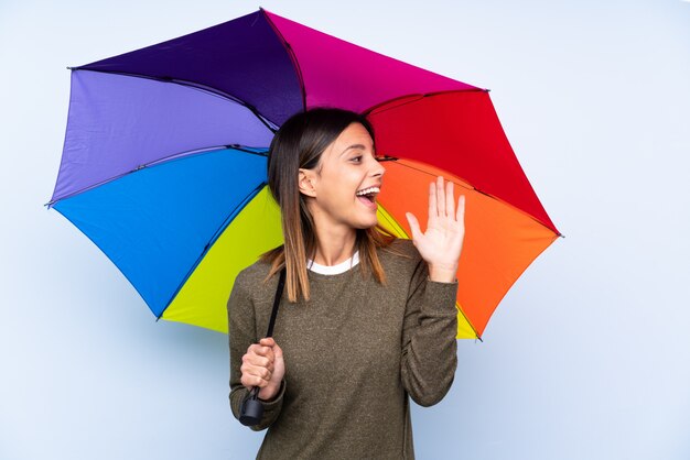 Junge brünette Frau, die einen Regenschirm über blauer Wand hält, die mit offenem Mund schreit