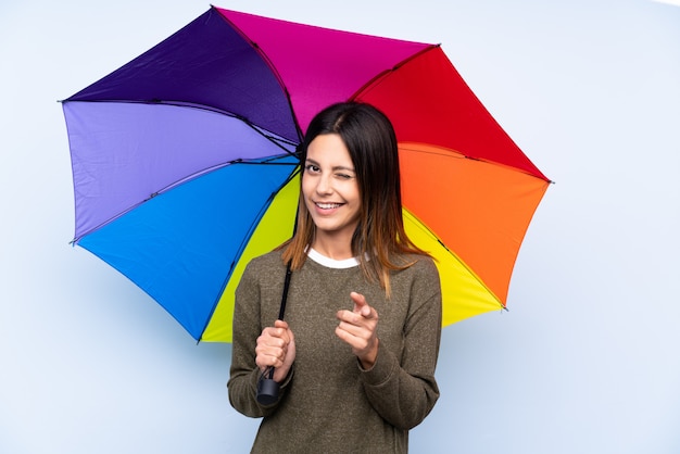 Junge brünette Frau, die einen Regenschirm über blauer Wand hält, zeigt Finger auf Sie