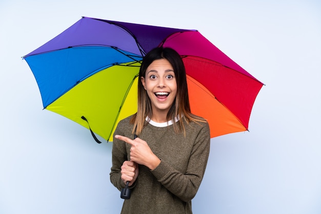 Junge brünette Frau, die einen Regenschirm über der blauen Wand hält, die Finger zur Seite zeigt