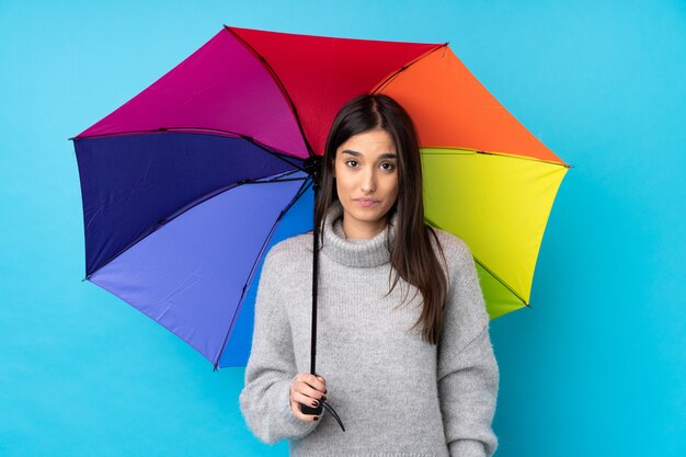 Junge brünette Frau, die einen Regenschirm über isolierte blaue Wand flehend hält