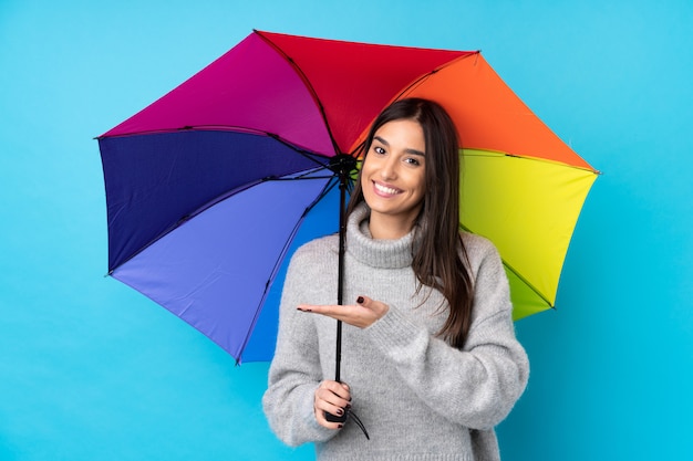 Junge brünette Frau, die einen Regenschirm über isolierter blauer Wand hält, die Hände zur Seite für Einladung zum Kommen ausdehnt