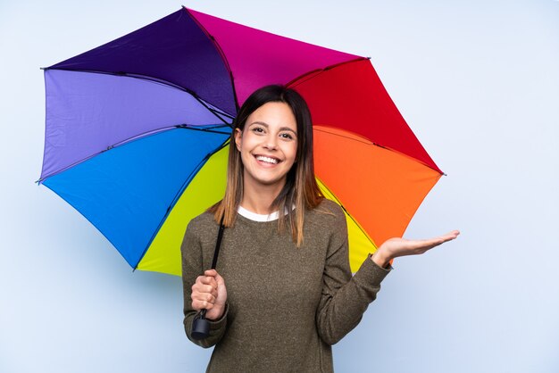 Junge brünette Frau, die einen Regenschirm über isolierter blauer Wand hält, die imaginären Copyspace auf der Handfläche hält