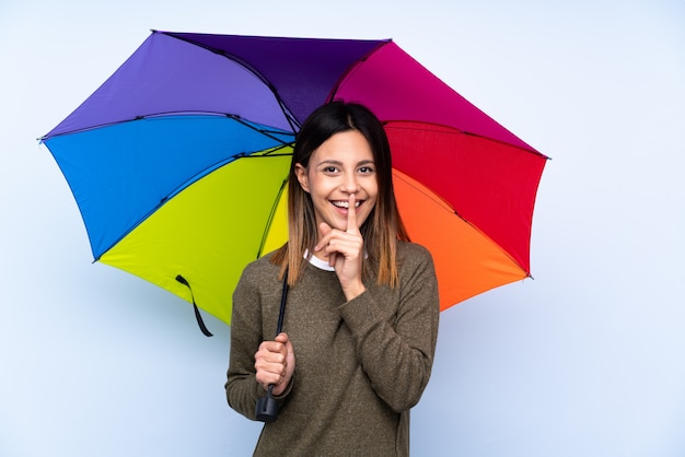 Junge brünette Frau, die einen Regenschirm über isolierter blauer Wand hält, die Schweigegeste tut