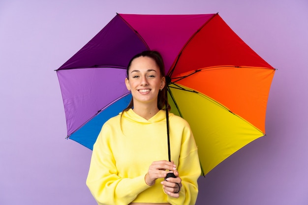 Junge brünette Frau, die einen Regenschirm über isolierter lila Wand hält, die viel lächelt