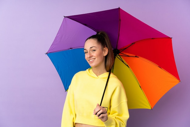 Junge brünette Frau, die einen Regenschirm über isolierter lila Wand hält, die viel lächelt