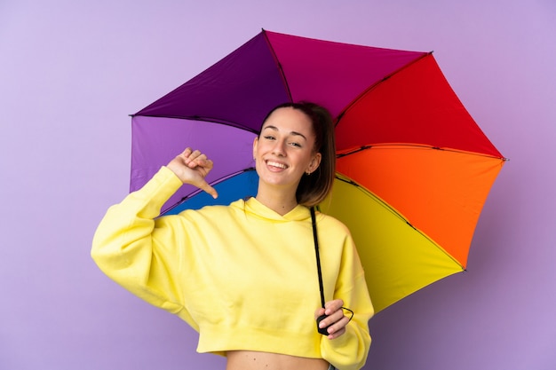 Junge brünette Frau, die einen Regenschirm über isolierter lila Wand hält, stolz und selbstzufrieden