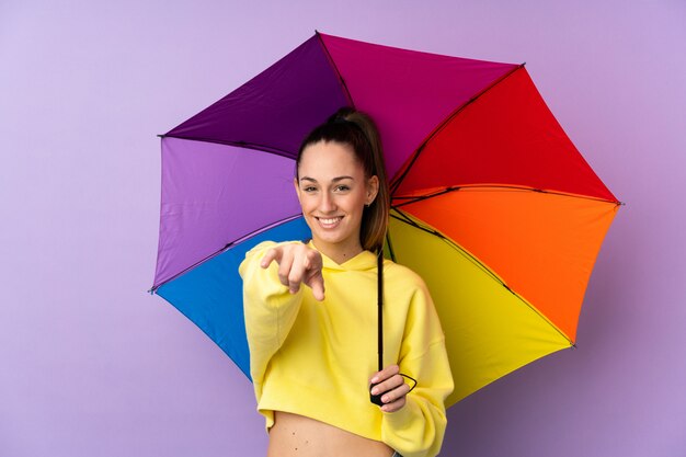 Junge brünette Frau, die einen Regenschirm über isolierter lila Wand hält, zeigt Finger auf Sie mit einem selbstbewussten Ausdruck