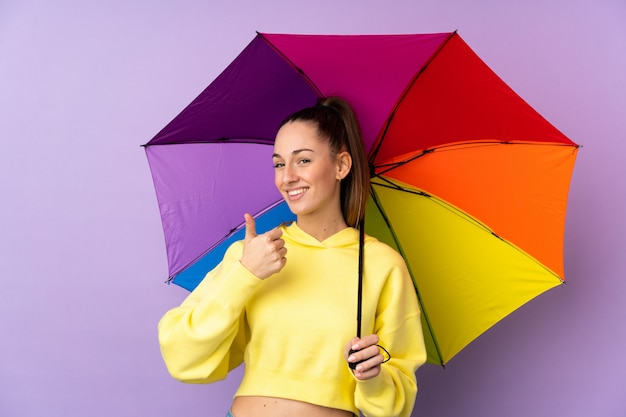 Junge brünette Frau, die einen Regenschirm über isolierter lila Wand mit Daumen hoch hält, weil etwas Gutes passiert ist