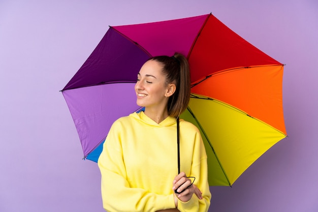 Junge brünette Frau, die einen Regenschirm über isolierter lila Wand mit glücklichem Ausdruck hält