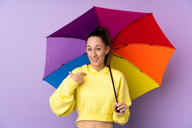 Junge brünette Frau, die einen Regenschirm über isolierter lila Wand mit überraschendem Gesichtsausdruck hält