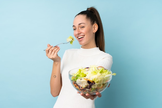 Junge brünette Frau, die einen Salat über isolierter blauer Wand hält